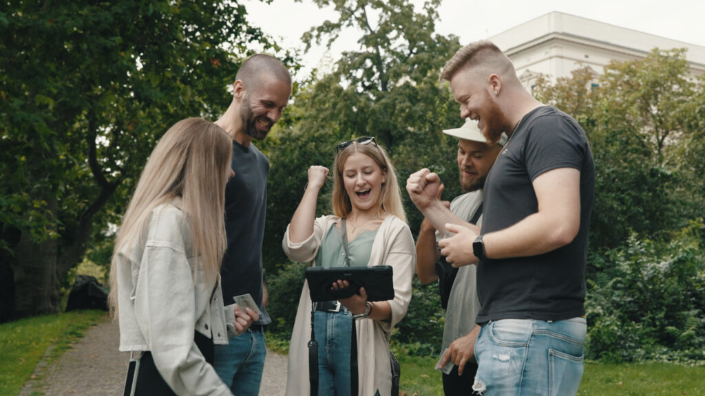 Gruppe von Freunden löst Outdoor Escape Game mit Tablet und Rätselkoffer im Park.