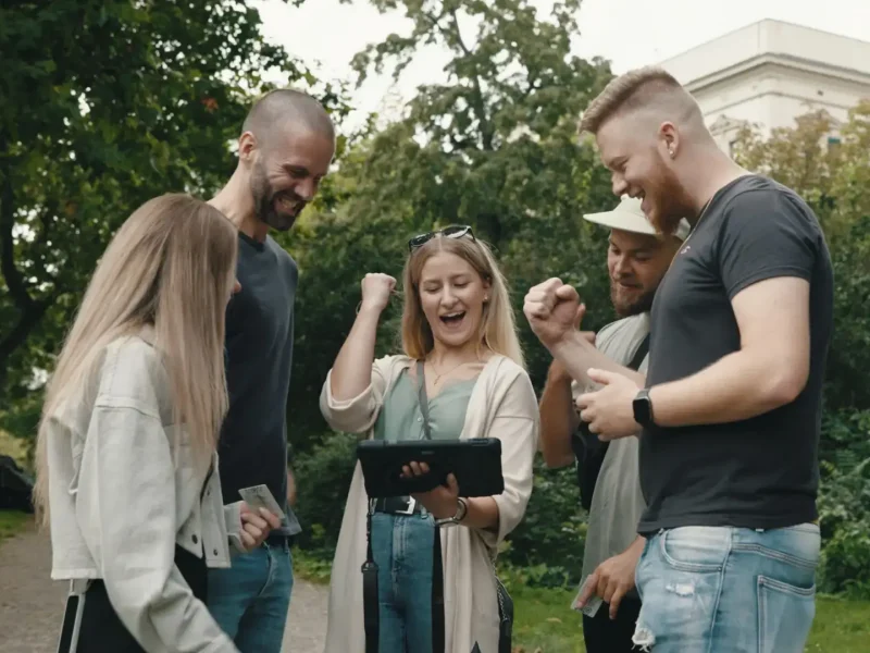 Gruppe von Freunden löst Outdoor Escape Game mit Tablet und Rätselkoffer im Park.