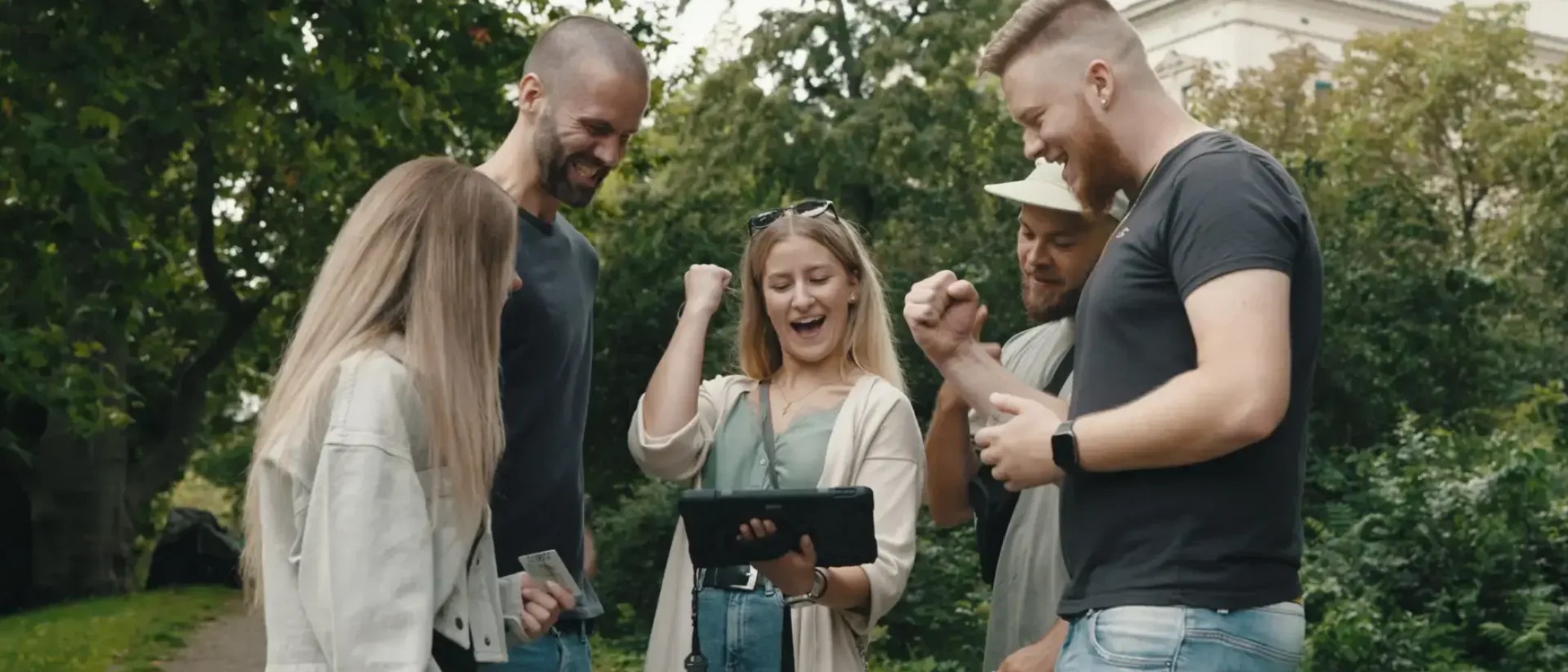 Gruppe von Freunden löst Outdoor Escape Game mit Tablet und Rätselkoffer im Park.